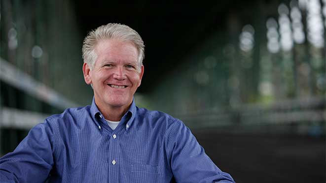 The author, in a seated position on a covered bridge. He is caucasian with blond-gray hair and blue eyes, wearing a royal blue long-sleeve shirt with thin, white, checkerboard pinstripes.