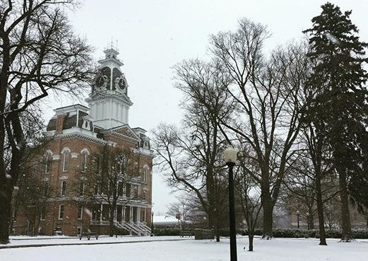 A brown and white college campus building set in a winter landscape with snow covering the ground, There are numerous deciduous trees scattered around which are bare of leaves, as well as an evergreen tree.