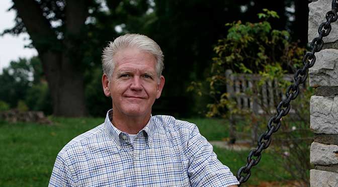A portrait of the author in a park-like setting, with dark green grass and various trees and bushes in the background. There is a stone pillar in the rith side of the photo with a heavy black chain coming from the top and draped down and to the left in front of the author, who has his left arm resting on it.