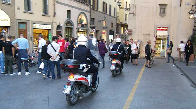 An active European street scene with two police scooters driving away from the viewer, numerous people walking and standing around between shops on either side of the street.