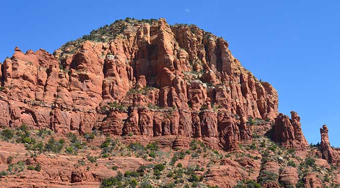 A desert scene with a large mountain in the center. The mountain is red in color, with layers of horizontal white stone in segments. The foreground is a reddish-colored sand floor with scrub plants and a deep blue sky overhead.