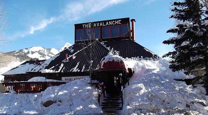 A frontier-style wooden saloon building with snow piled halfway up all sides of the walls. The stairs leading up to the front door have been dug out, The sky is clear blue with a wispy cloud above the rooftop, and a snow-covered evergreen to the right of the front stairs.