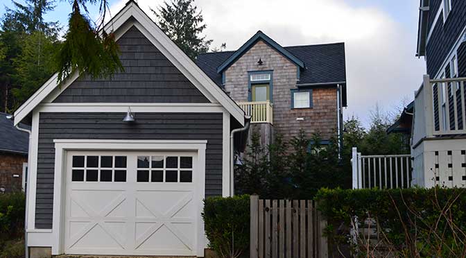 A personal residence with a dark gray two-car garage on the left with white trim, A wooden fence extends off the right side of the garage and continues to the right where it adjoins a two story house painted with the same colors. There is another two story house with wooden siding and blue trim located at the back of the property. A line of small trees separated the garage and read house.