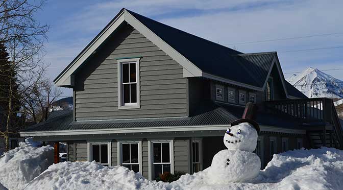A gray two-story home set in a winter landscape. Snow is piled up in front of the house with a snowman in the right, front foreground, mixed foliage trees on the left and a snow-covered mountain in the background with blue sky overhead.