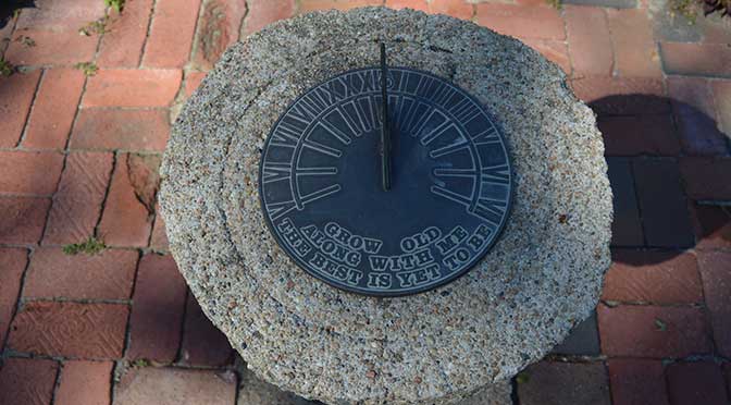 An outdoor stone Sundial, with aged bronze face, is set against a brick walkway background.