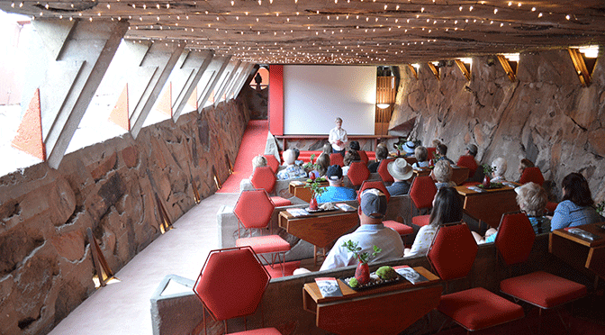 A classroom setting viewed from the back of the room. An instructor is standing at the front of the room with about a dozen older adults seated in chair facing her. Light is coming in from open windows on the left side of the room, illuminating the red furniture upholstery and stone walls of the interior.