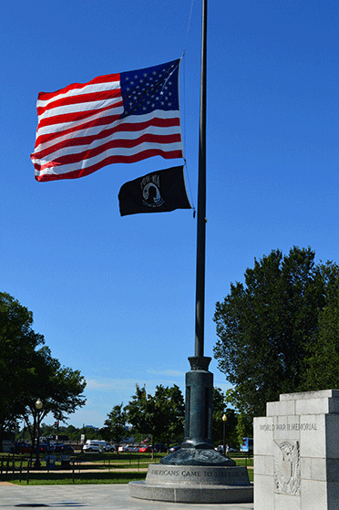 The United States flag flies at half mast, with the PoW flag beneath it. There is a clear blue shy overhead, and scattered desert plants in the background.