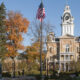 Hillsdale College campus during autumn, with an American flag displayed on a common area in front of a multi-storied brown brick building with white trim. The courtyard in the foreground shows green grass, numerous trees in various autumn colors and a clear, deep blue shy overhead.